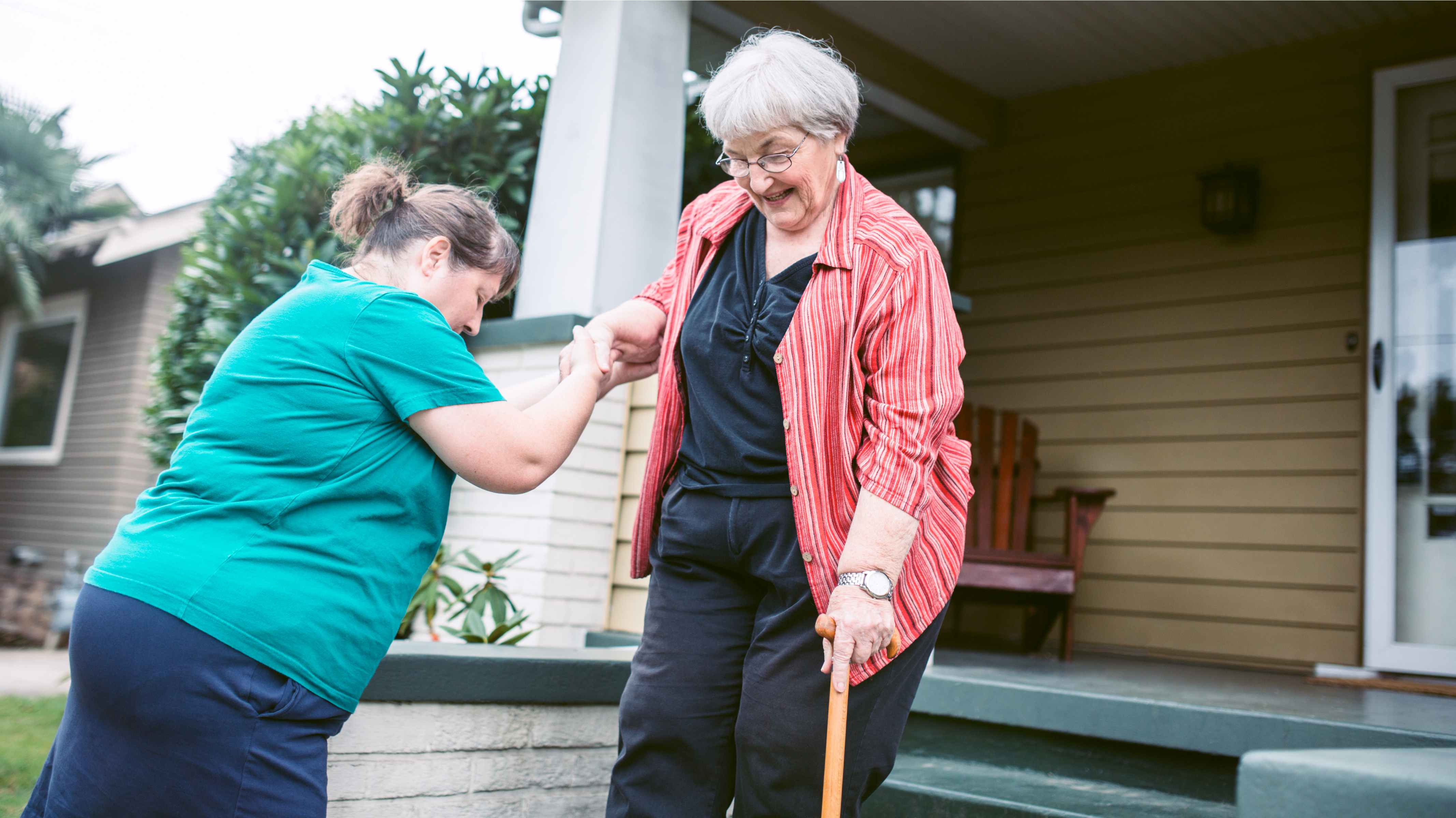 A woman assists a senior citizen with a cane climb down some stairs outside a house