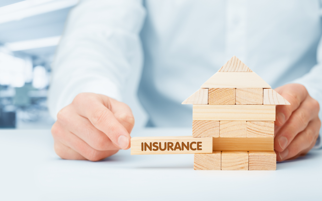 A person holds a house made of wooden blocks and points at block that reads "insurance". 